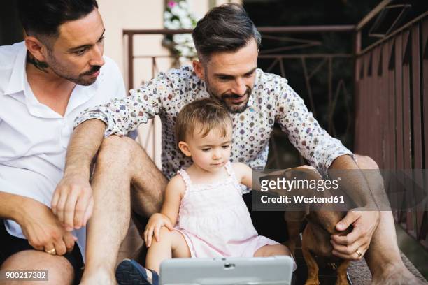 Gay couple with daughter and dog on balcony using digital tablet
