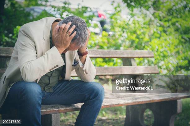 man with head in his hands sitting on park bench - manos a la cabeza fotografías e imágenes de stock