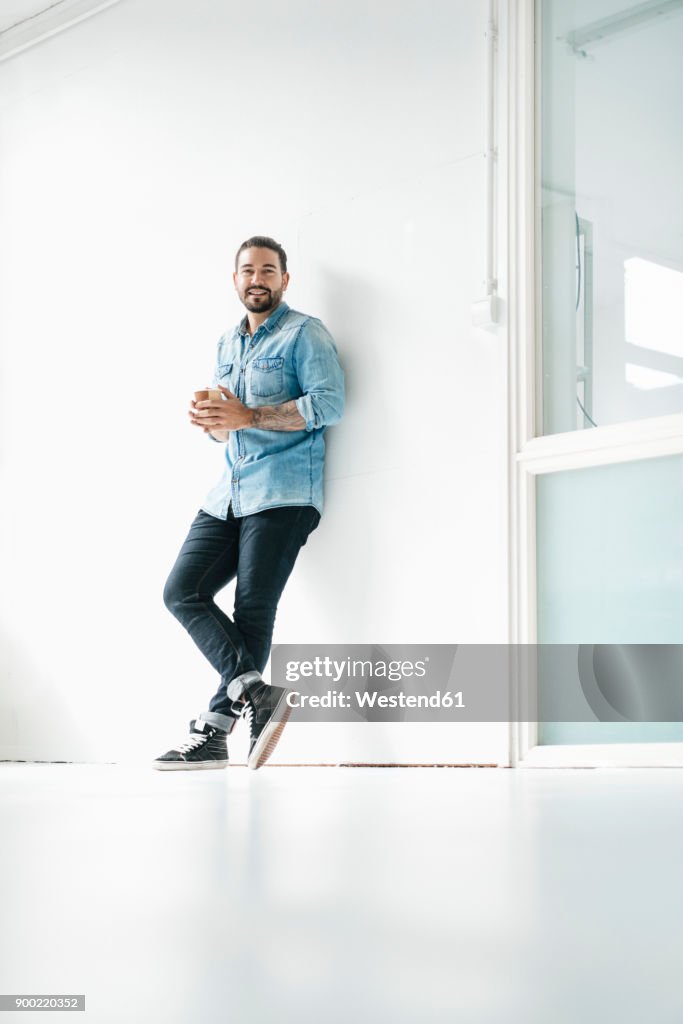Portrait of smiling man with coffee to go leaning against wall in a loft