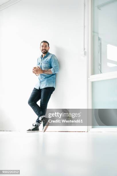 portrait of smiling man with coffee to go leaning against wall in a loft - lehnend stock-fotos und bilder