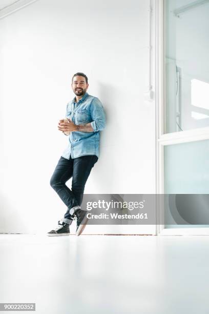 portrait of smiling man with coffee to go leaning against wall in a loft - anlehnen stock-fotos und bilder