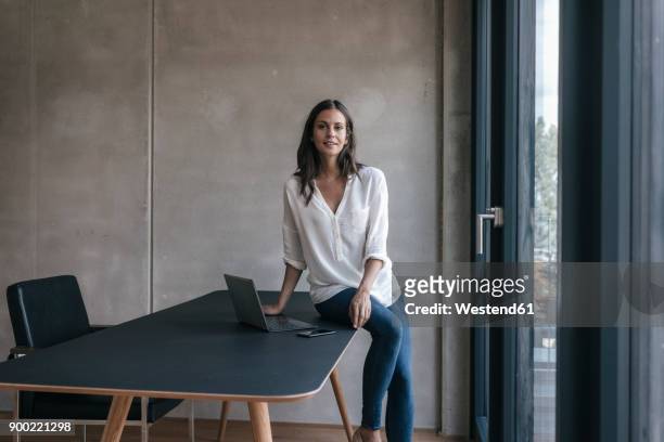 portrait of smiling woman sitting on table with laptop and cell phone - lässige kleidung stock-fotos und bilder