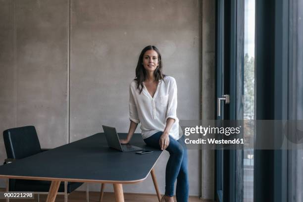 portrait of smiling woman sitting on table with laptop and cell phone - business woman sitting imagens e fotografias de stock