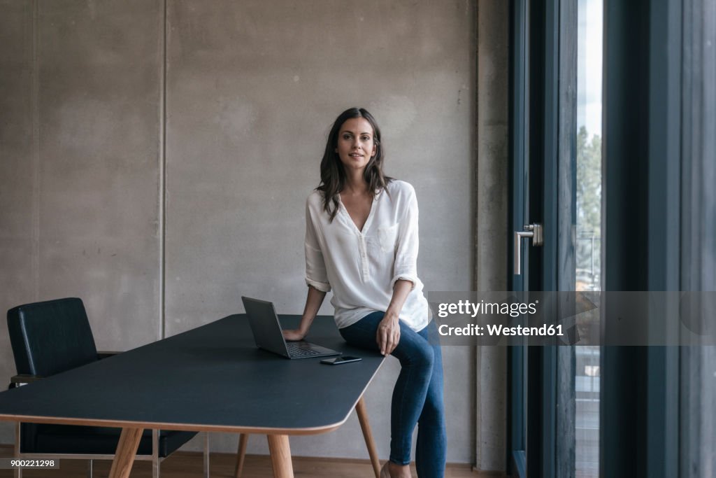 Portrait of smiling woman sitting on table with laptop and cell phone