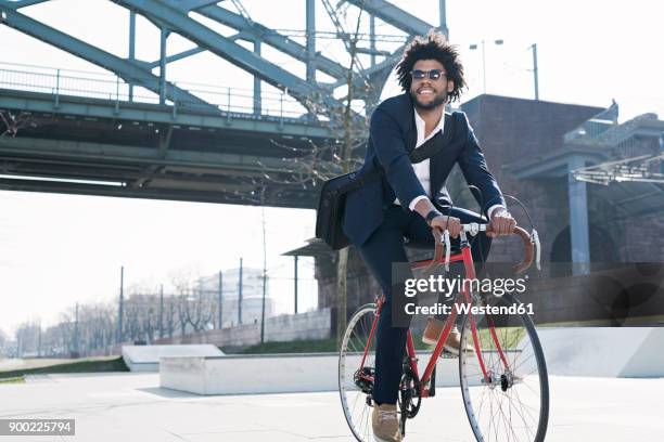 smiling businessman riding bicycle at riverside bridge - black suit sunglasses stock pictures, royalty-free photos & images