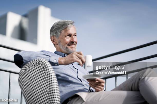 smiling grey-haired man sitting on balcony and drinking coffee - gelassene person stock-fotos und bilder