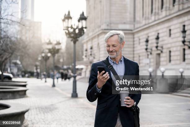 grey-haired businessman in the city with smartphone and coffee to go - man on cell phone walking in the city stock pictures, royalty-free photos & images
