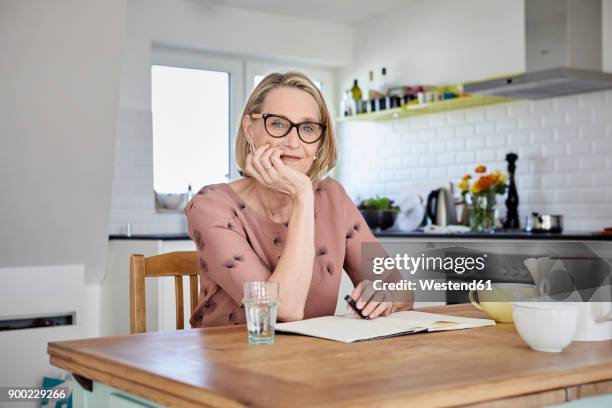 portrait of mature woman with notebook at kitchen table - 50s woman writing at table imagens e fotografias de stock