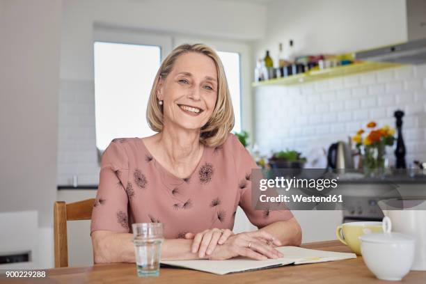 portrait of smiling mature woman with notebook at kitchen table - 50s woman writing at table imagens e fotografias de stock
