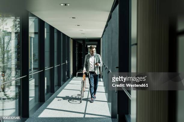Businessman with cell phone pushing bicycle in office passageway