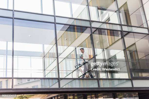 businessman with cell phone pushing bicycle in office passageway - paso elevado fotografías e imágenes de stock