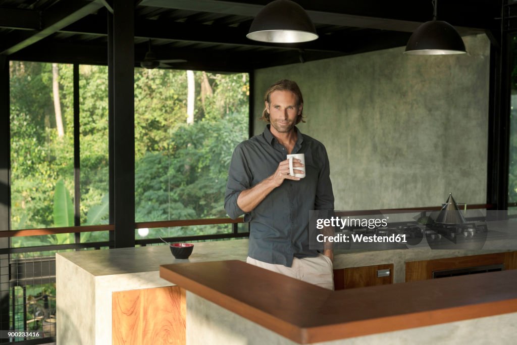 Handsome man drinking coffee in modern design kitchen with glass facade surrounded by lush tropical garden