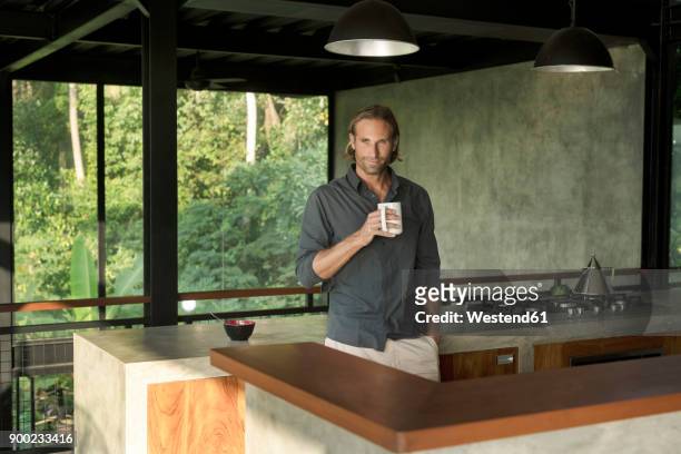 handsome man drinking coffee in modern design kitchen with glass facade surrounded by lush tropical garden - luxury kitchen stock-fotos und bilder