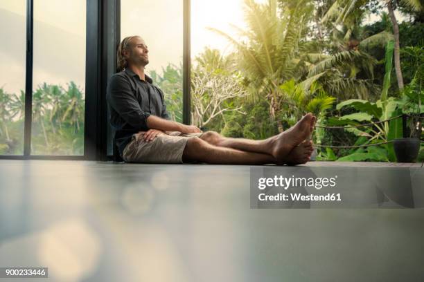 handsome man sitting on floor and leaning on glass facade with stunning tropical garden in background - bare feet male tree stock pictures, royalty-free photos & images