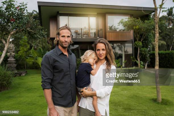 portrait of smiling family standing in front of their design house surrounded by lush tropical garden - two parents stock-fotos und bilder