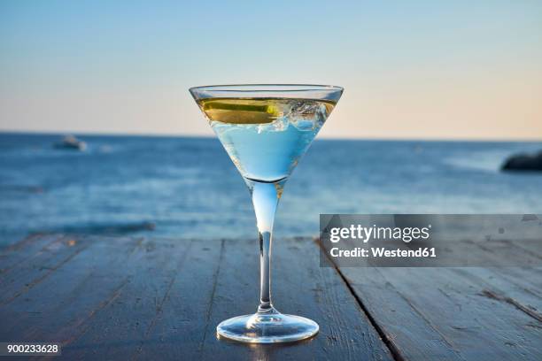 glass of ice-cooled martini with lime slice in front of the sea - martini glass stock-fotos und bilder