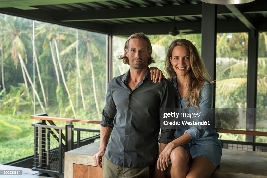 Couple smiling at camera in contemporary design house with glass facade surrounded by lush tropical garden