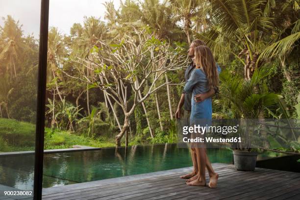 hugging couple standing on wooden terrace in front of pool and enjoying stunning view in lush tropical garden - poolside stock pictures, royalty-free photos & images