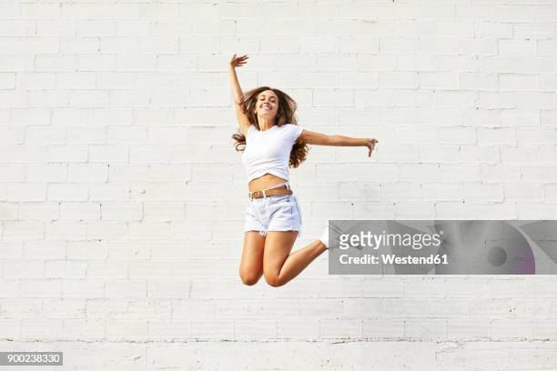 happy young woman jumping mid-air in front of white wall - women in daisy dukes stock pictures, royalty-free photos & images