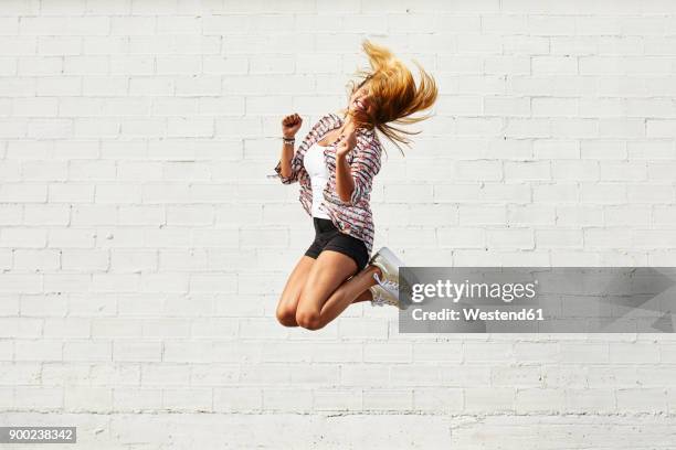 happy young woman jumping mid-air in front of white wall - calções coloridos imagens e fotografias de stock
