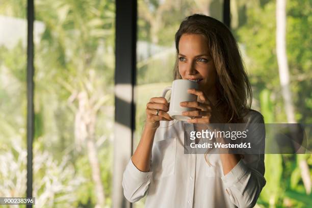 smiling woman drinking coffee at home in front of lush tropical garden - cafe front photos et images de collection