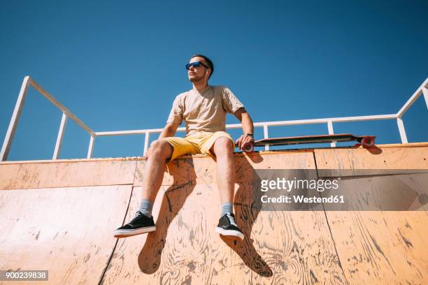 young man with earbuds and longboard sitting on top of halfpipe in skatepark - skateboarding half pipe stock pictures, royalty-free photos & images
