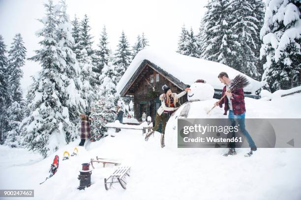 austria, altenmarkt-zauchensee, friends building up big snowman at wooden house - winter hut stock pictures, royalty-free photos & images