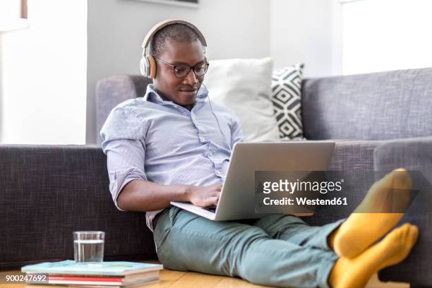 young man sitting on the floor in the living room using laptop and headphones - tutorial stock-fotos und bilder