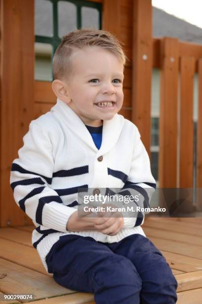 toddler boy fake cheesy smile; sitting on a wooden play set - fake smile stock-fotos und bilder