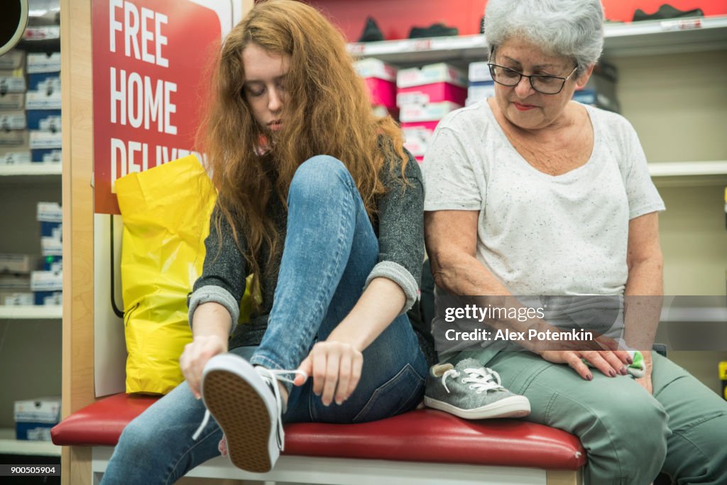 The silver-haired 65-years-old active senior woman and her unhappy teenager granddaughter shopping shoes in the clothing retail store