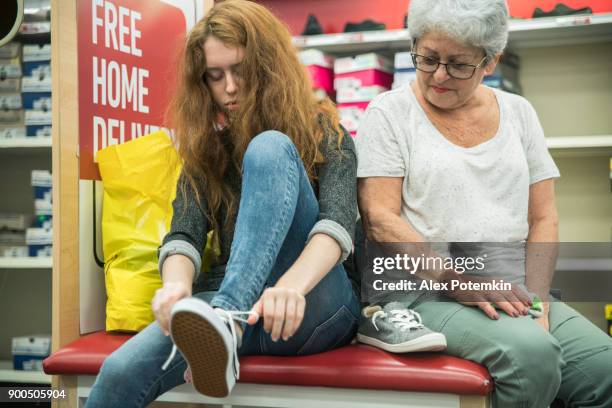 the silver-haired 65-years-old active senior woman and her unhappy teenager granddaughter shopping shoes in the clothing retail store - shopping disappointment stock pictures, royalty-free photos & images