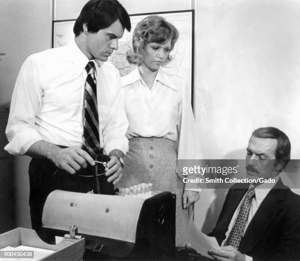 Publicity still showing three actors, Robert Urich, Maureen Reagan and Jack Hogan, posing on set, in an office/lab with a bag full of test tubes,...