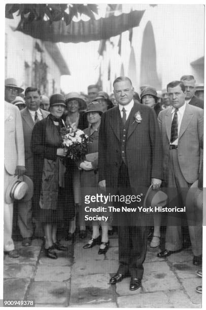 In front of crowd, President Herbert Hoover stands, hat in hand, with his wife, First Lady Lou Henry Hoover, in a cloche hat, who holds a bouquet,...