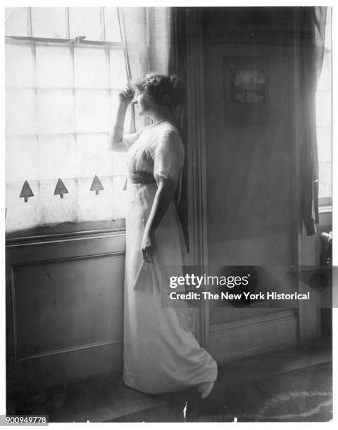 Actress Frances Maule standing at a window, holding a fan in her left hand, right hand pulling back curtain, 1922.