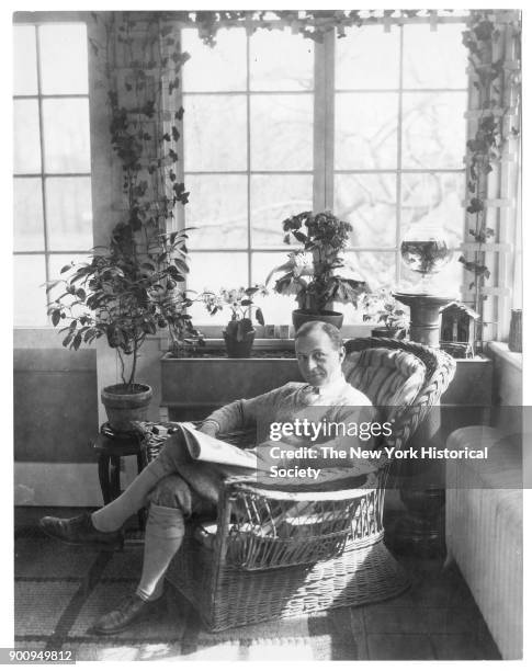 Actor Ernest Truex, in sweater and knee-high socks, seated in wicker armchair with magazine or newspaper on lap, before window with plants on sill,...