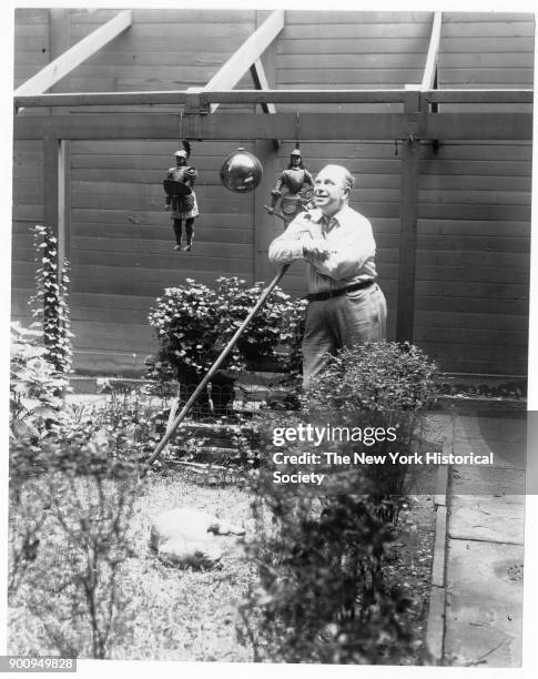 Puppeteer Tony Sarg, leaning on hoe in garden, at 52 W 9th Street, looking toward sky, cat at his feet, 1939.