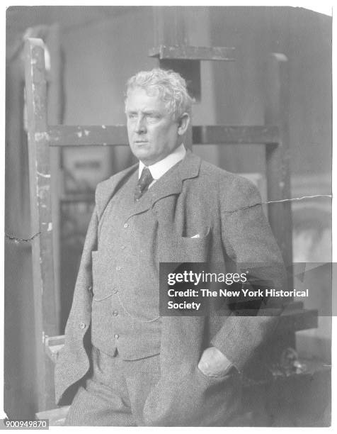 Painter J Alden Weir, in three-piece tweed suit with hands in pockets, standing in front of an empty easel, 1912.