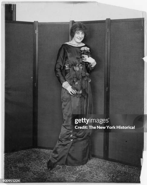 Model standing before screen in floor length gown with lace color and cuffs; holding stem of flowers, 1911.