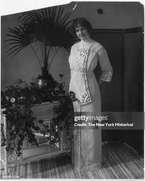 Model in floor-length dress with embroidery on front placket; standing next to wicker stand with ivy, flowers, a potted palm, 1911.