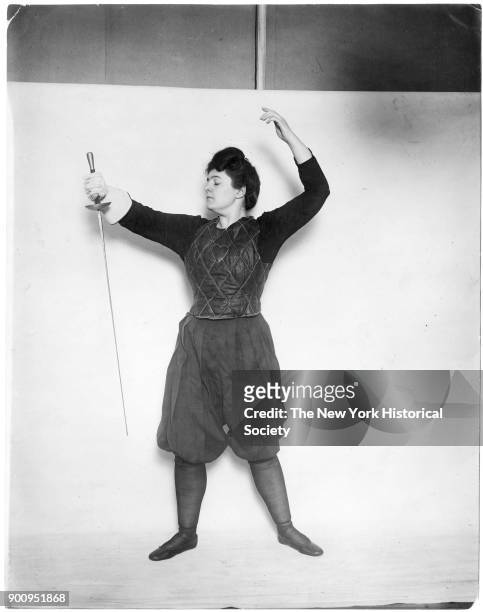 Model in front of backdrop, dressed in fencing outfit with heart applique on chest, holding sword, 1911.