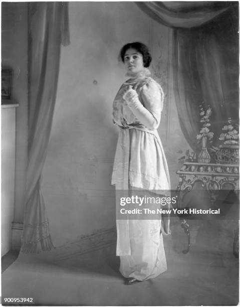 Model in front of painted backdrop, wearing floor length draped light dress, arm raised to her shoulder, 1911.
