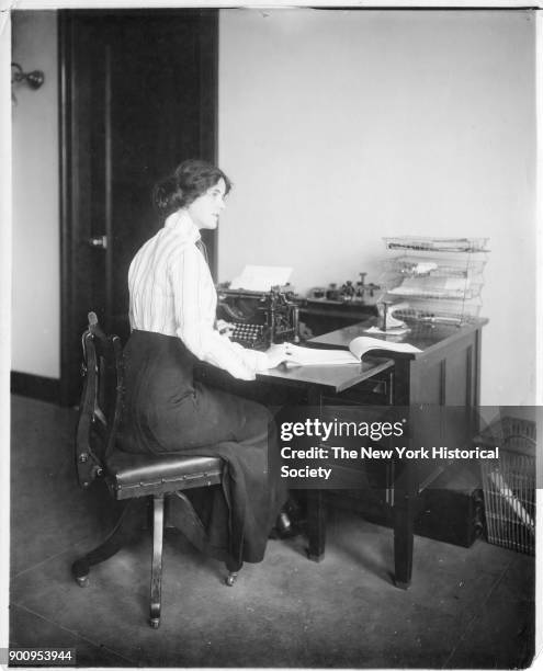 Model seated at desk with typewriter and tablet of paper, wearing pinstriped shirt and dark, high-waisted skirt, 1911.