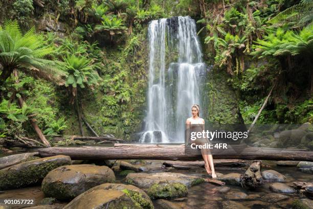 mooie vrouw zitten op een log in de voorkant een prachtige waterval, terug naar de natuur, beauchamp falls, great ocean road - redwood stockfoto's en -beelden