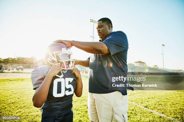 football coach helping young player put on helmet before practice - american football strip stock pictures, royalty-free photos & images