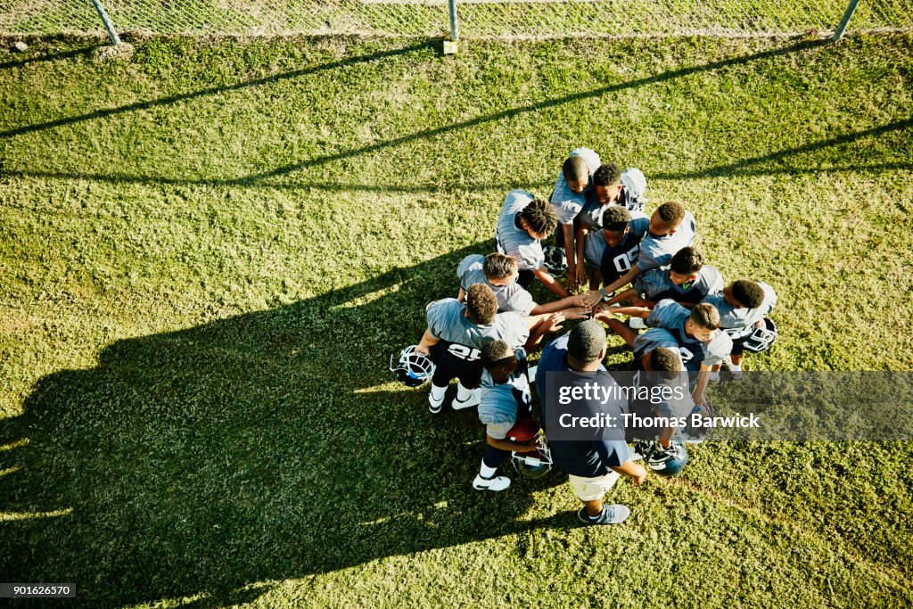 Overhead view of coach and football team gathered in circle with hands together before football game