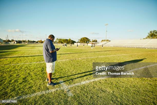 football coach working on smartphone during practice with young football players - innocence project stock pictures, royalty-free photos & images