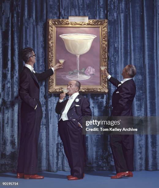 Portrait of three men of differing heights, all dressed in tuxedos and all drinking margaritas underneath a large painting of a margarita mounted...