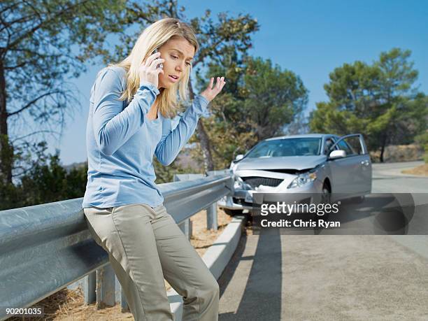 frustrated woman using cell phone next to car wrecked on guardrail - car accident stock pictures, royalty-free photos & images