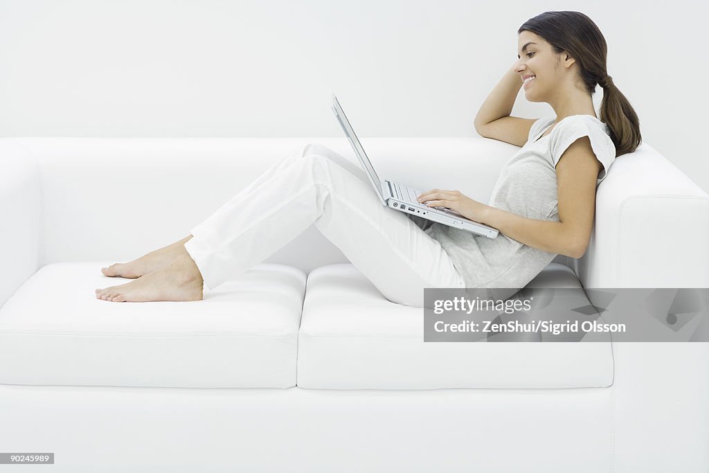 Teenage girl reclining on couch, using laptop, side view