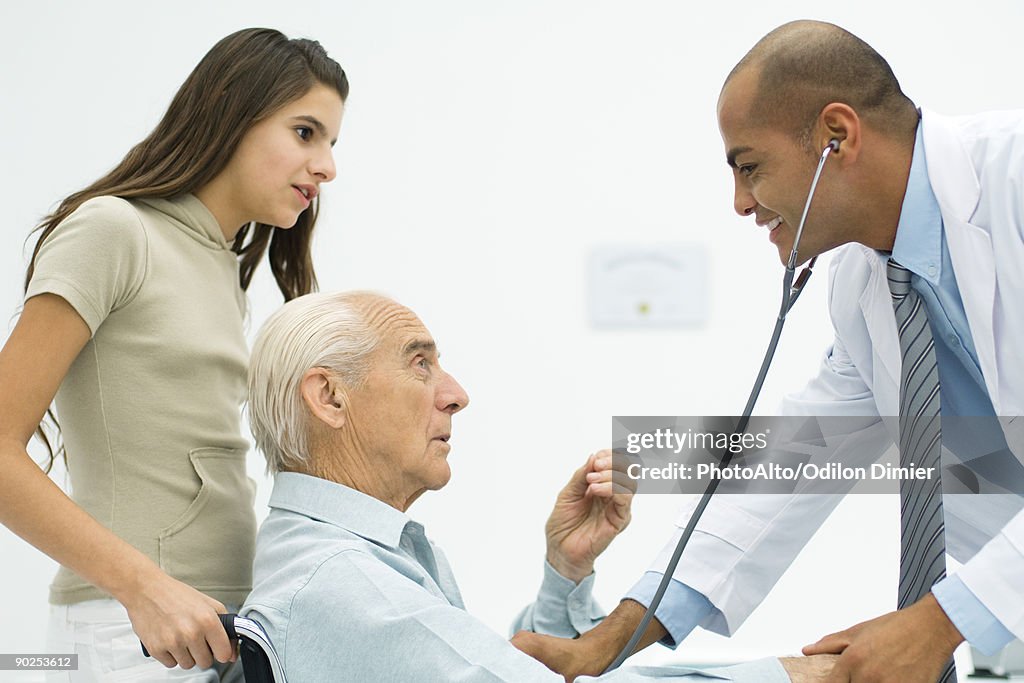 Doctor listening to senior man's heart, teen girl watching